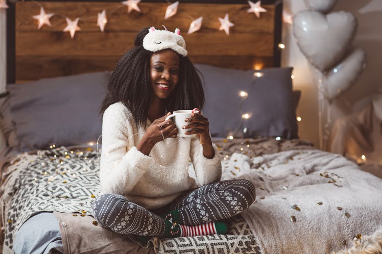 A happy woman in festive pjs holds a coffee mug on her bed with string lights in the background.