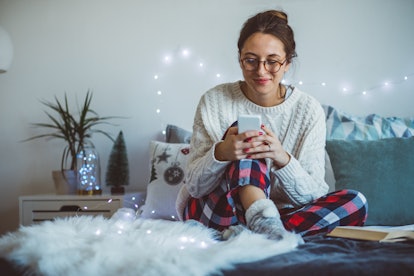 A woman in Christmas loungewear types on her phone, while sitting in her bed. 