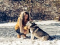 A happy woman kneels next to her husky in the snow.