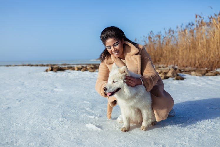 A happy woman in a pea coat poses with her white husky in the snow on a sunny day.