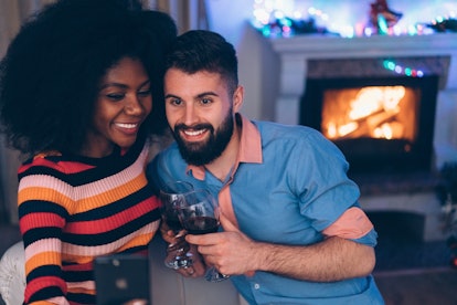 A couple takes a selfie with wine glasses next to their fireplace in the winter. 