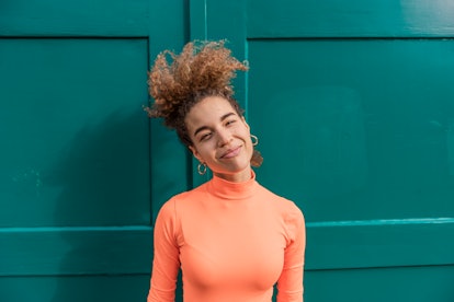 A woman in an orange turtleneck stands in front of a green garage. 