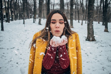 A woman in a yellow coat and red sweater blows on a snowball in the winter. 
