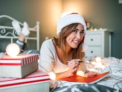 A happy woman writes out a Christmas card on her bed.