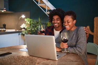 A happy couple holds red wine glasses while looking at their laptop at home.