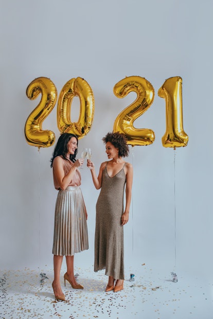 Two friends clink their glasses while standing next to gold, 2021 balloons on New Year's Eve.
