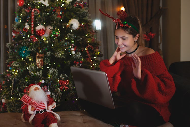 A woman dressed up for holidays with reindeer ears, sits at her laptop, next to her Christmas tree.