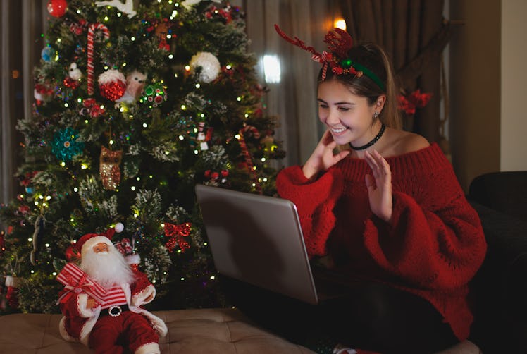 A woman dressed up for holidays with reindeer ears, sits at her laptop, next to her Christmas tree. 