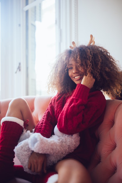A young Black woman poses in a Christmas sweater and socks while attending a virtual Christmas party...