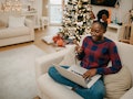 A young Black woman sits in her decorated living room with a laptop and attends a virtual Christmas ...