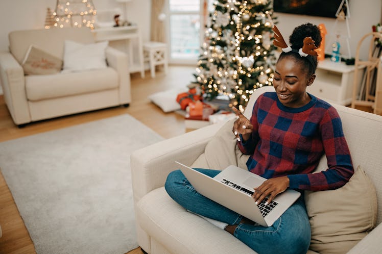 A young Black woman sits in her decorated living room with a laptop and attends a virtual Christmas ...