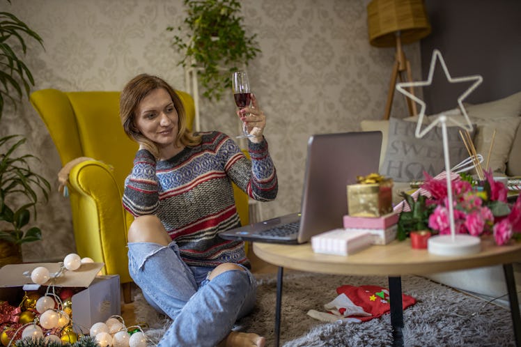 A woman holds up her glass of red wine while looking at her laptop in her festively-decorated living...