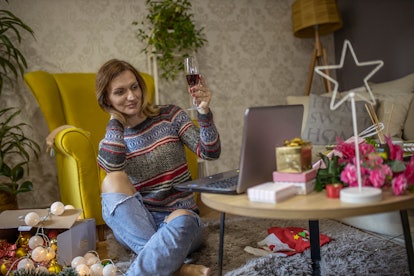 A woman holds up her glass of red wine while looking at her laptop in her festively-decorated living...