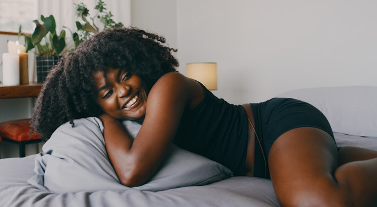 A young Black woman lays in bed and smiles while wearing loungewear.