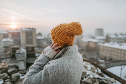A young woman wearing a yellow winter hat and gray jacket stands on a rooftop and looks over a snowy...