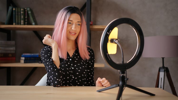 A happy woman with a black and white blouse and pink hair takes a selfie using a ring light and phon...