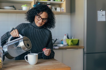 A woman with a gray sweater on pours herself tea in her kitchen.