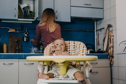 A baby sits in a yellow high chair eating while a woman stands with her back facing the camera at a ...