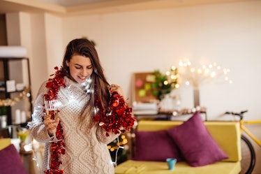 A woman is festive and cozy at home with fairy lights and a red garland draped around her neck, whil...