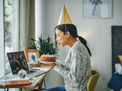 A young woman blows out candles on a cake, while attending a virtual birthday party for adults and w...