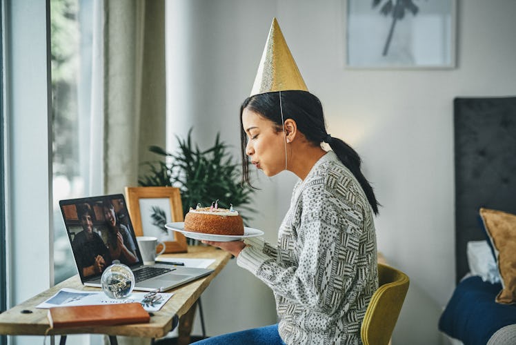 A young woman blows out candles on a cake, while attending a virtual birthday party for adults and w...