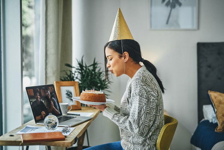 A young woman blows out candles on a cake, while attending a virtual birthday party for adults and w...