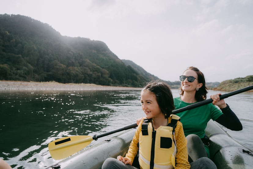 A woman and a young girl kayak on a river among rolling green hills.