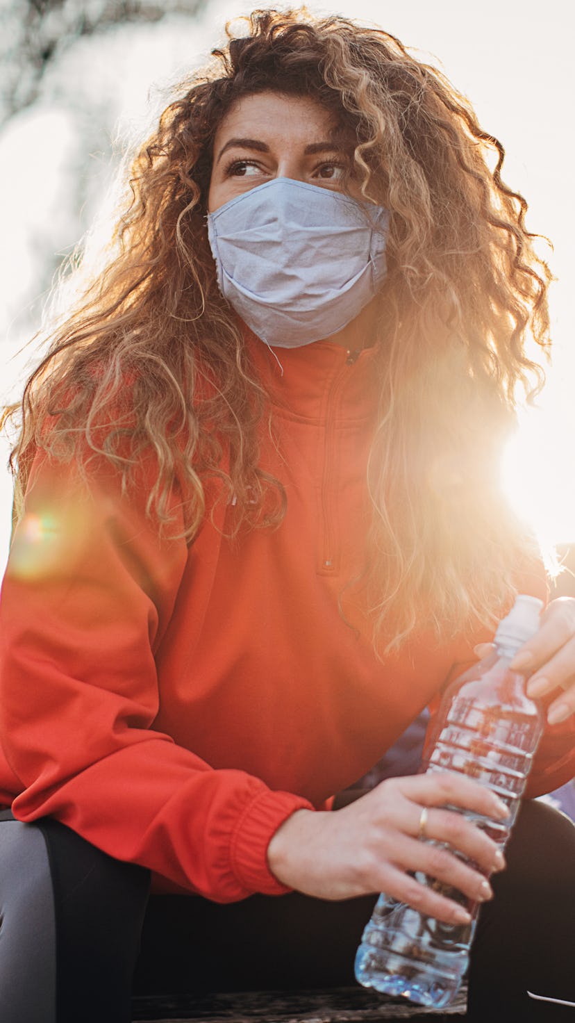 A person with natural hair wears a mask and holds a water bottle while sitting on a park bench. Work...