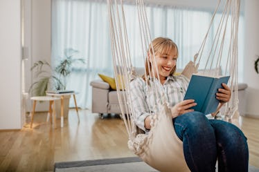 A young woman sits in a crocheted swing in her living room and reads a book on a cozy Hanukkah night...