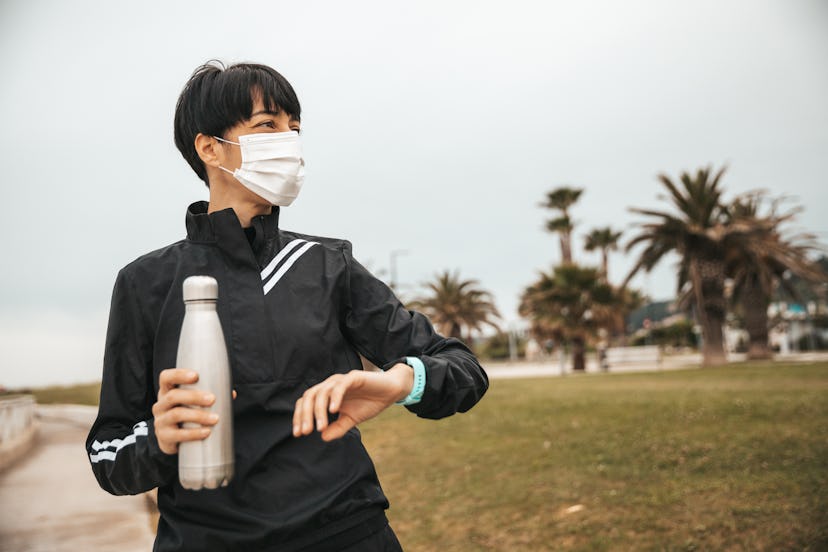 A person holds a water bottle and checks their watch while out for a run wearing a mask. Exercise is...