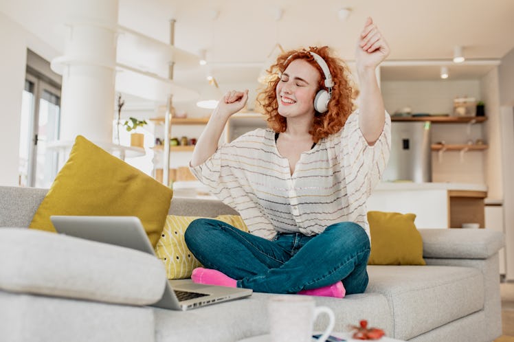 A happy woman throws up her hands on her couch while sitting next to her laptop.
