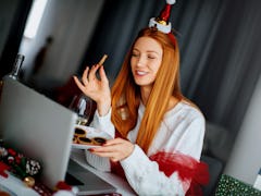 A happy woman wearing a Santa hat holds a plate of Christmas cookies while chatting with friends on ...