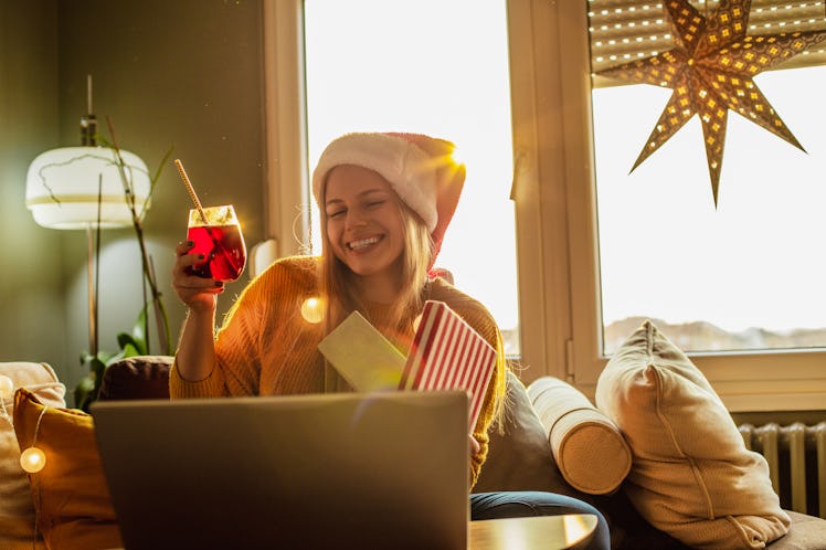 A young woman smiles while holding a present and participating in virtual Secret Santa with her best...