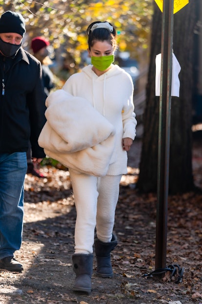 Selena Gomez in a cream tracksuit and black Ugg boots, with a lime face mask, carrying a cream plush...