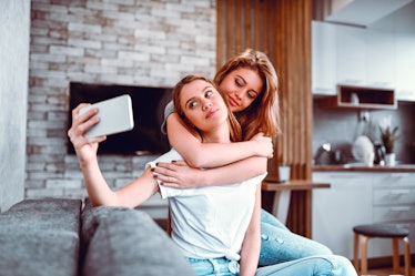 A couple hugs and takes a selfie on their phone, while sitting in the living room at home. 