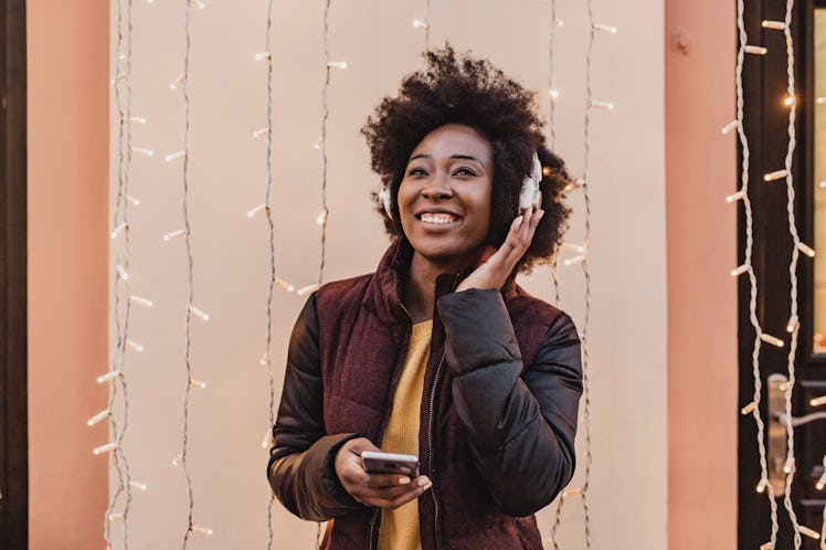 A young Black woman smiles in front of strands of Christmas lights while listening to holiday music ...