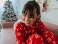 A happy woman in Christmas pajamas sits on her couch, next to her Christmas tree. 