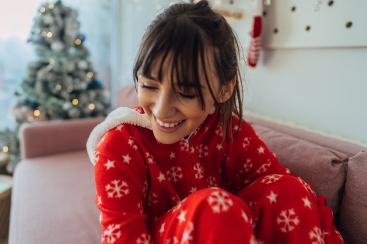 A happy woman in Christmas pajamas sits on her couch, next to her Christmas tree. 