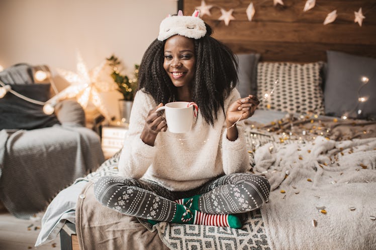 A woman in Christmas pajamas sits on her bed with twinkle lights and a mug of hot cocoa. 
