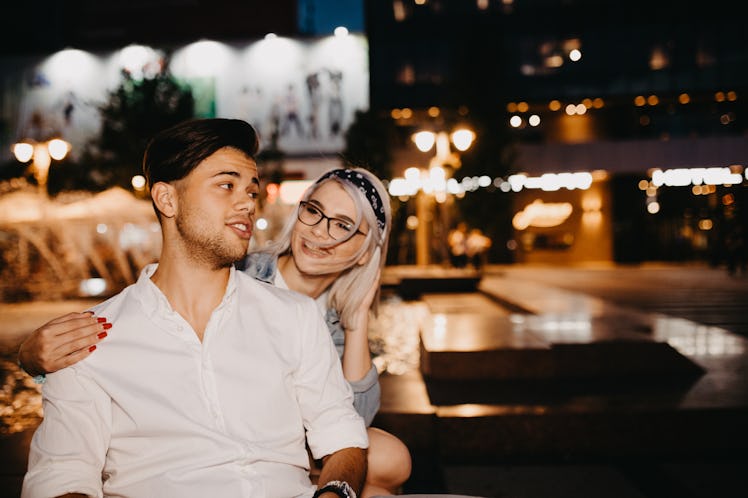 A couple hangs out in the city together, with bright lights glowing in the background.