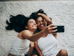 A couple laughs and takes a selfie while laying on a white carpet on the floor.