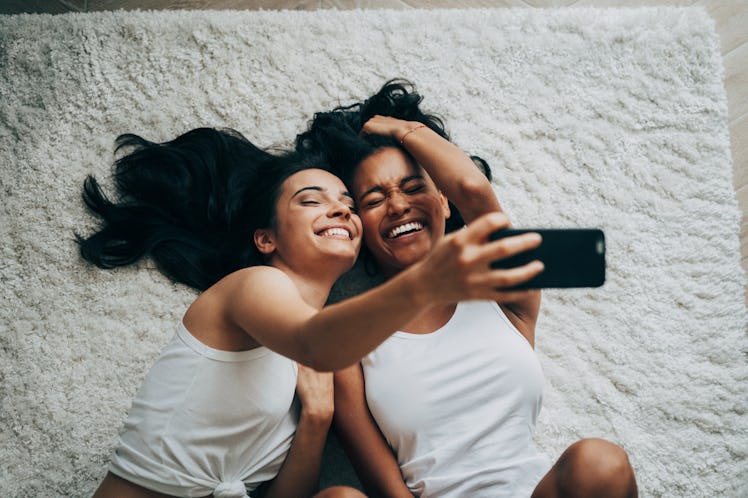 A couple laughs and takes a selfie while laying on a white carpet on the floor.