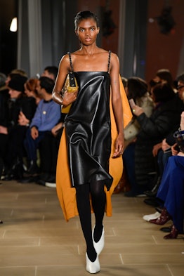 A model in a black leather dress and an orange cape shifting to reveal more of the model’s right sid...