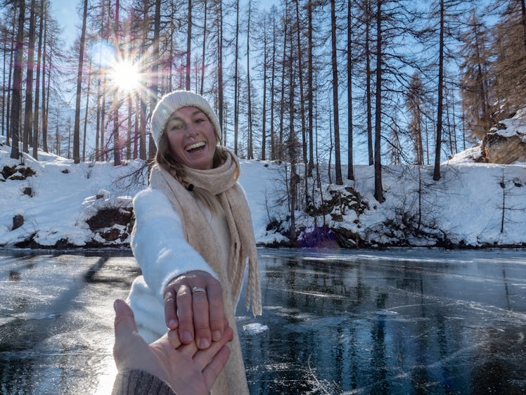 A happy women wearing a scarf, sweater, and beanie skates on an icy pond while holding her partner's...