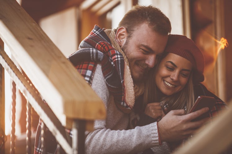 A happy couple cuddles up under a blanket on the steps while looking at a cellphone.