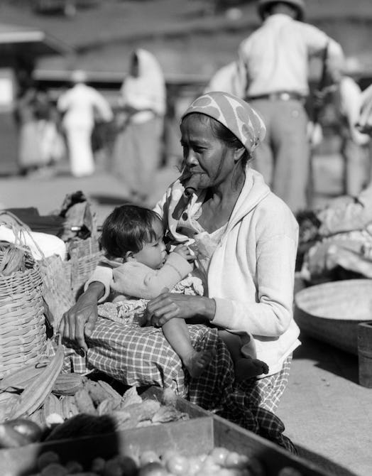 1920s 1930s WOMAN MOTHER SITTING SMOKING CIGAR CHEROOT HOLDING BABY CHILD IN MARKET PLACE BAGUIO PHI...