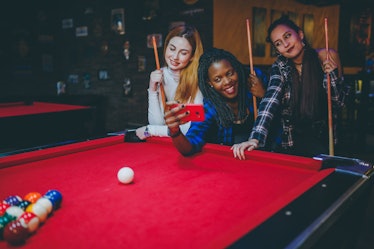 Three friends take a selfie on their phone while playing pool in a bar.