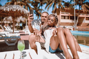 A couple relaxes on a lounge chair in the sunshine at a tropical beach resort while posing for a sel...