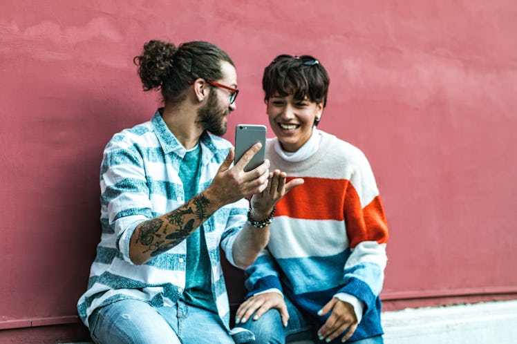 A couple sits against a pink wall in the city and laughs while looking at a phone.