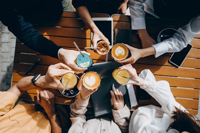 A group of friends holds their cups of coffee together at a coffee shop on a sunny day.